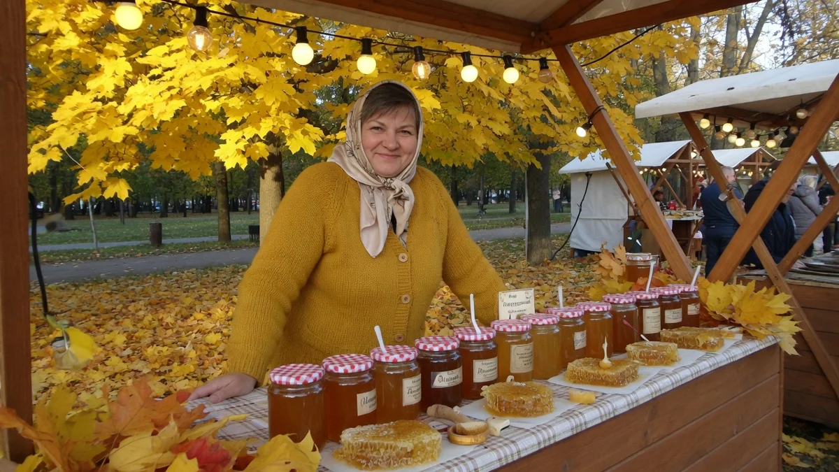 Продавщица мёда в горчичном кардигане за прилавком с банками на осенней ярмарке