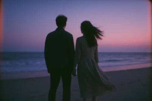 A couple stands on the shore holding hands against the backdrop of the sunset