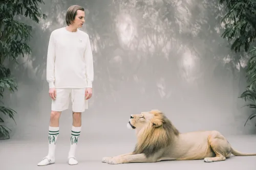 A young man in white clothing stands next to a relaxed lion
