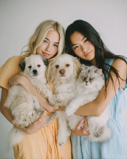 Dos chicas con perros lindos y un gato frente a una pared neutral