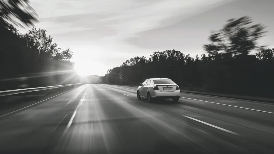 A light-colored car moving down a road against a sunset backdrop with trees
