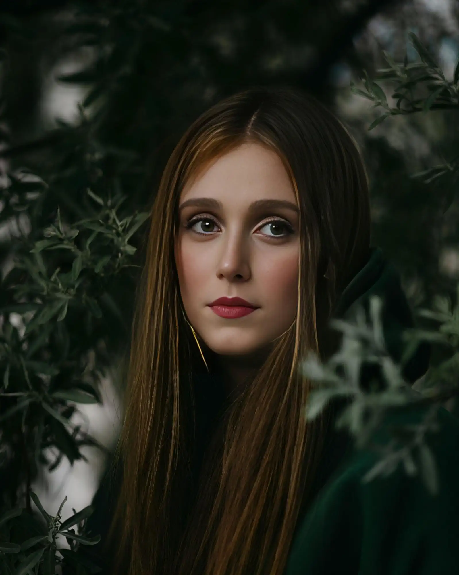 Portrait of a young woman with long light-brown hair and expressive gray-green eyes; she stands among dense dark foliage, soft light highlighting her even skin tone and nude makeup.