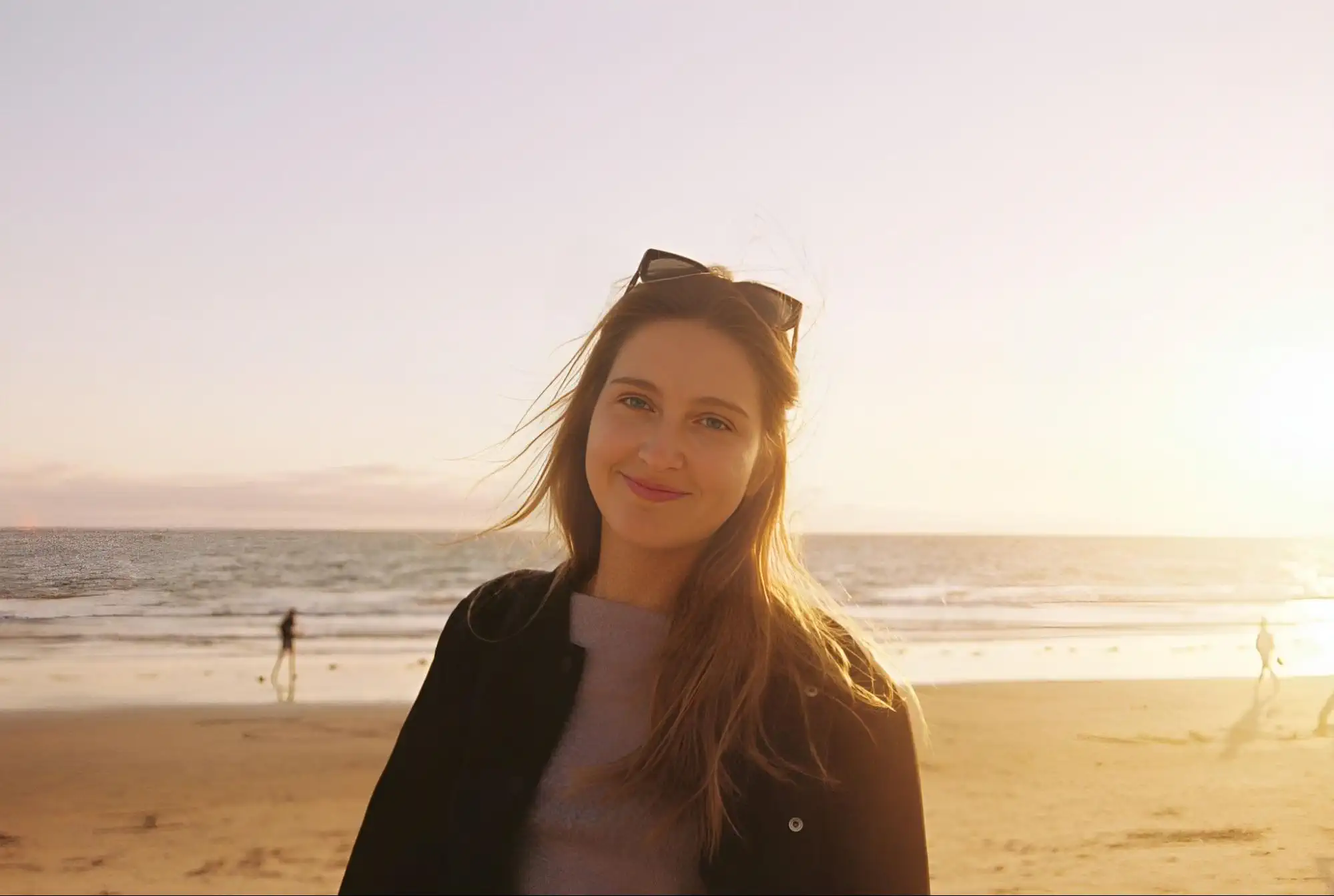A smiling girl on a sandy beach at sunset: the sun behind her lights her face with a warm glow, wind blows through her hair, she wears sunglasses, and waves and strolling silhouettes are visible in the distance.