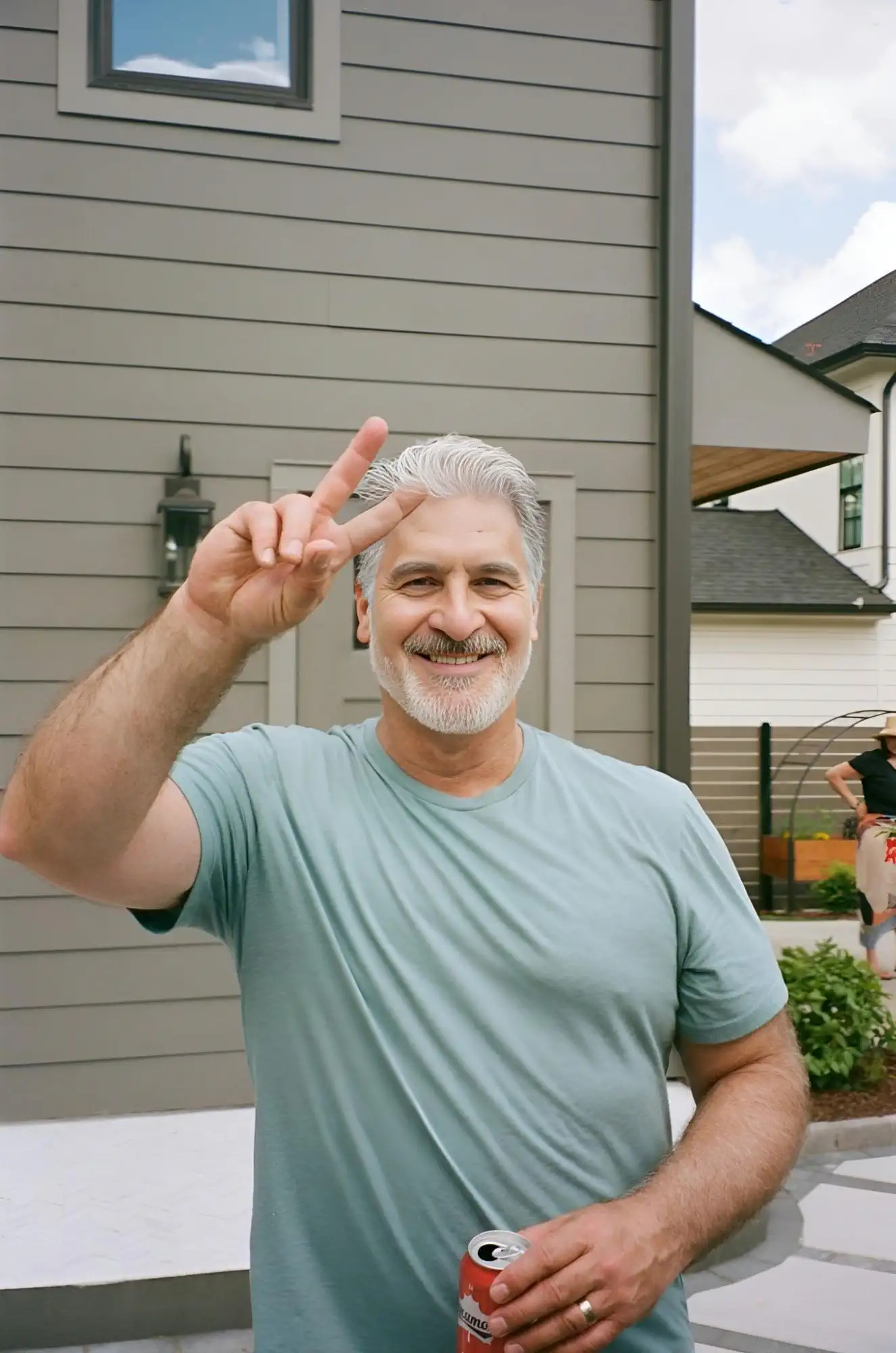 A gray-haired man in a blue T-shirt smiles and flashes a peace sign; in his other hand he holds a red drink can, with the facade of a modern house and a yard behind him.