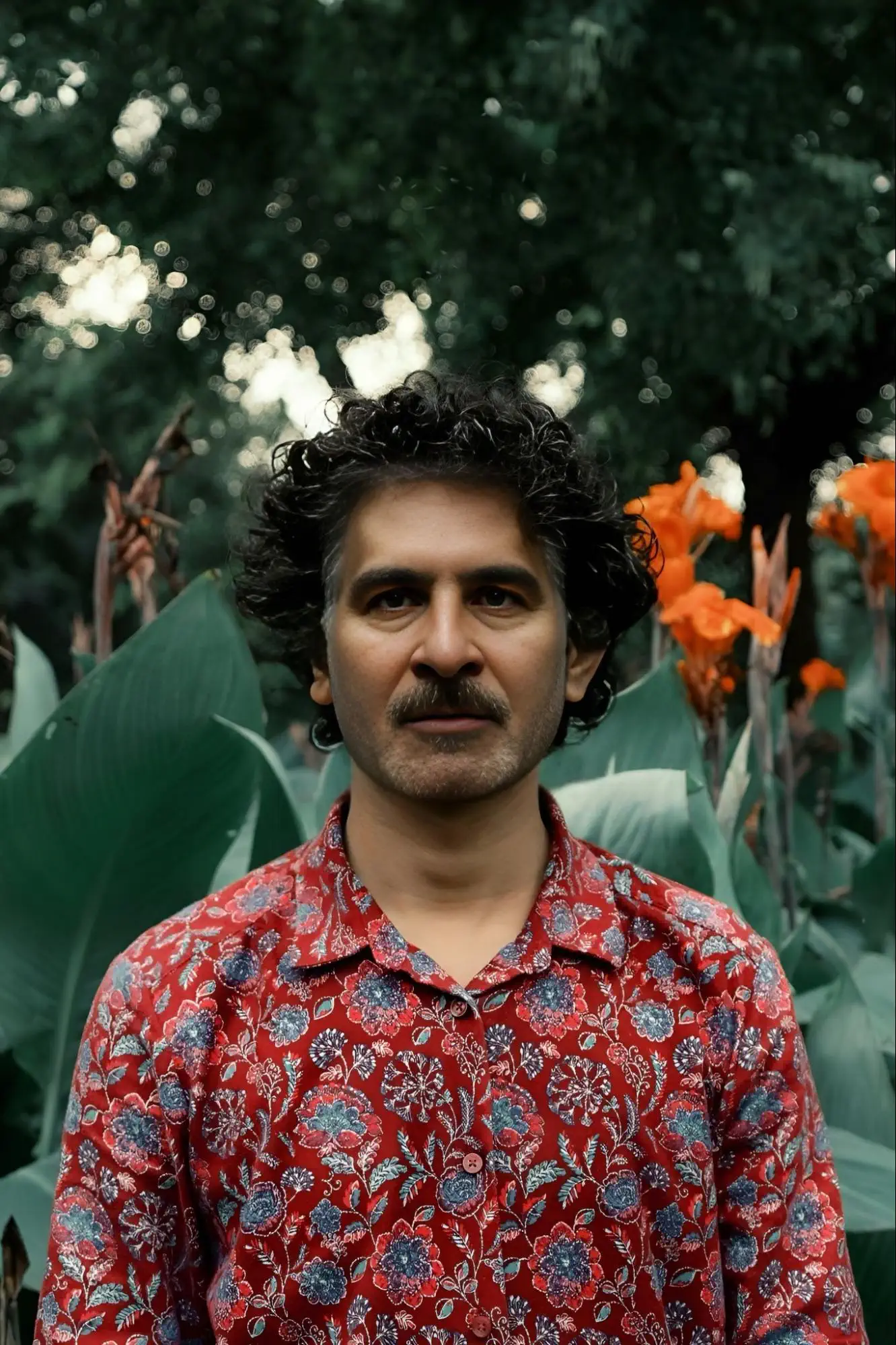Portrait of a man with dark curly hair and a mustache; he stands in front of lush green foliage and bright orange flowers, wearing a red shirt with a blue-turquoise floral pattern.