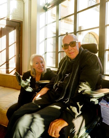 Два человека общаются в помещении; один из них в инвалидной коляске возле окна.