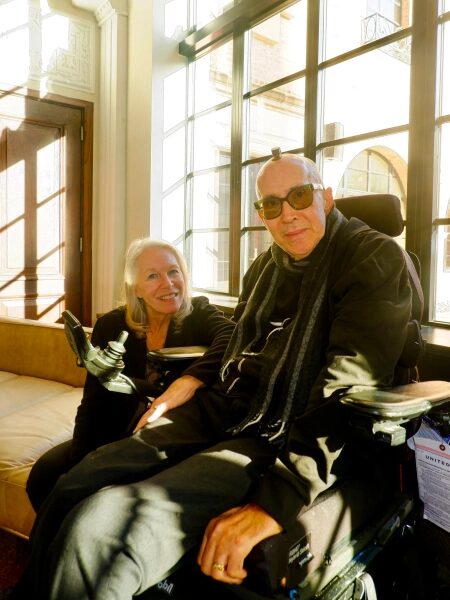 Два человека общаются в помещении; один из них в инвалидной коляске возле окна.