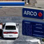 Человек заправляет белую машину на бензоколонке ARCO, рядом ценник на бензин.