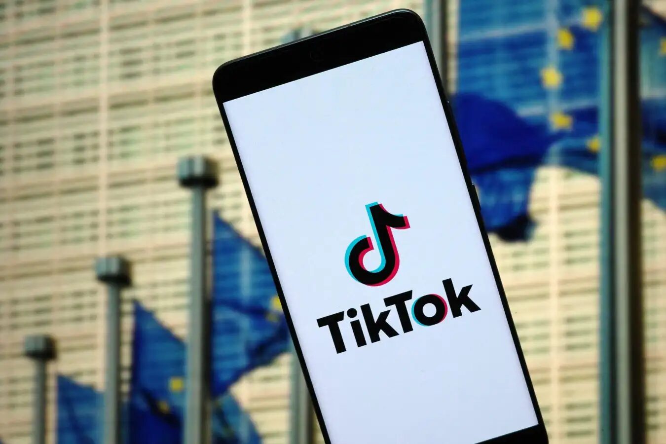 Нераскрытая реклама в TikTok обходит запрет на профилирование несовершеннолетних. 3 Смартфон с логотипом TikTok на фоне здания и флагов Европейского союза.