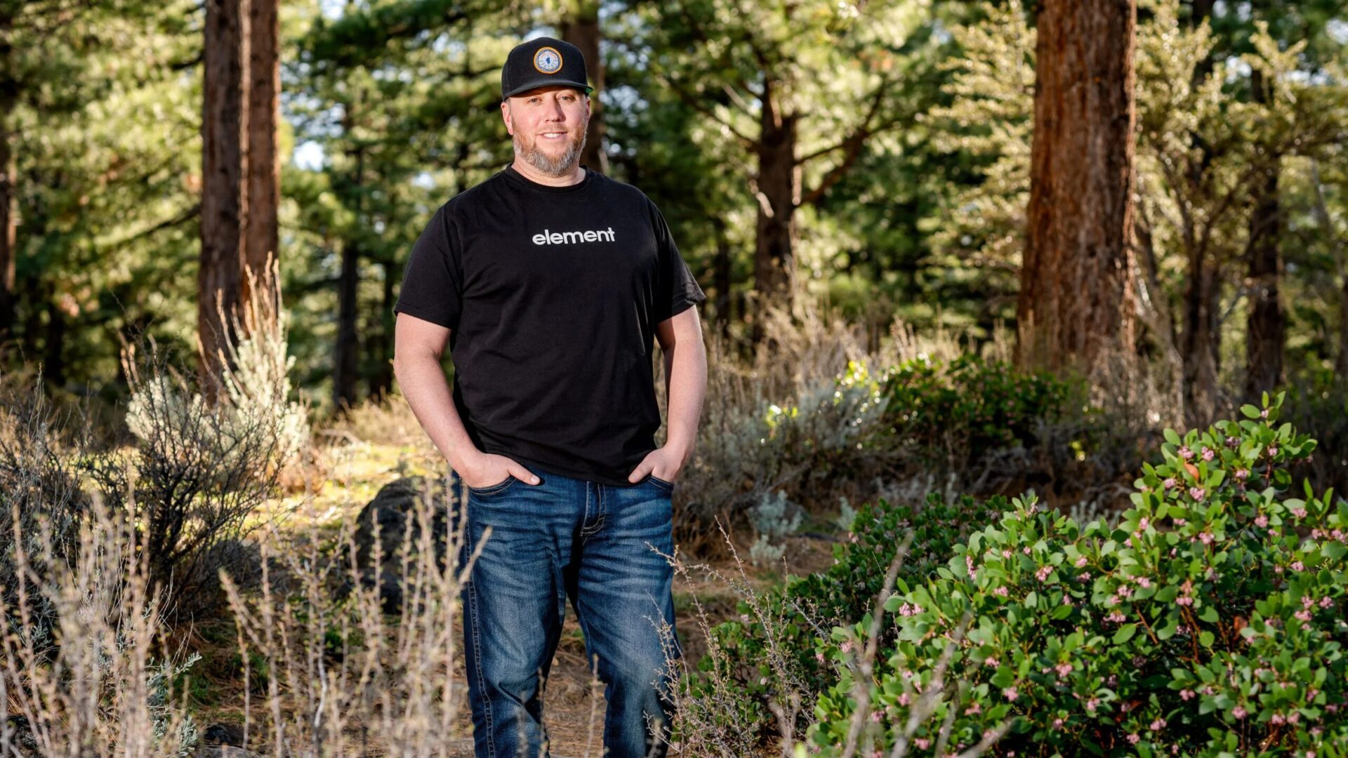 Мужчина в черной футболке и джинсах стоит среди леса.