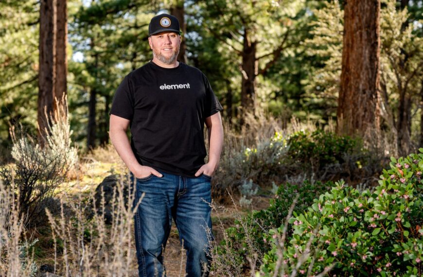 Человек в черной футболке и джинсах стоит на фоне леса.