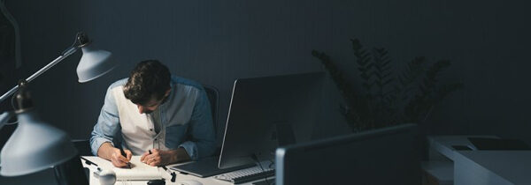 Человек работает за компьютером ночью, освещая рабочее место настольной лампой.