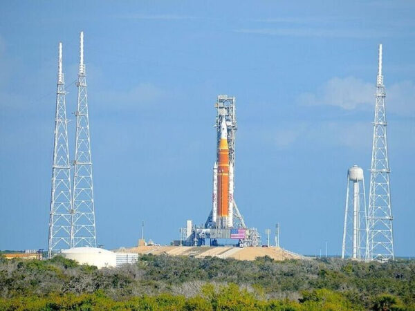 Ракета на стартовой площадке на фоне ясного неба, окруженная башнями и зеленью.