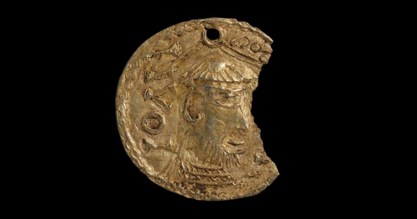 Золотое украшение с профилем мужчины, поврежденное с одной стороны, на чёрном фоне.