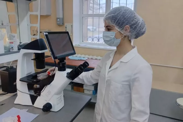 Женщина в лаборатории работает с микроскопом, одетая в белый халат и защитную шапочку.