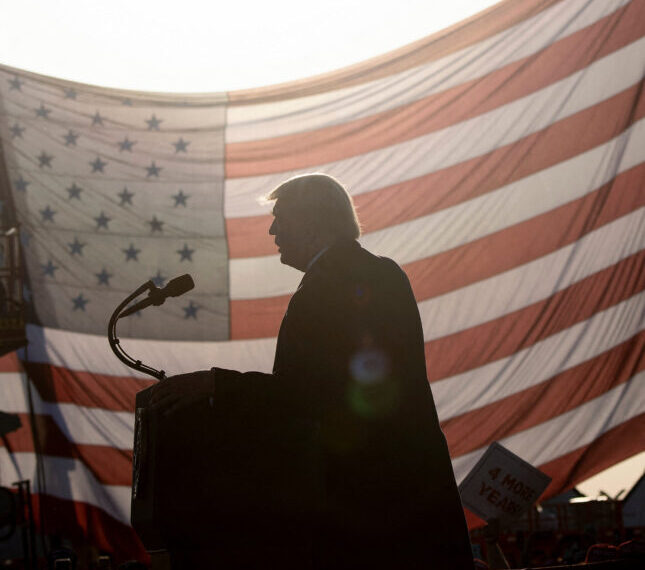 Политик за трибуной на фоне большого американского флага на митинге.