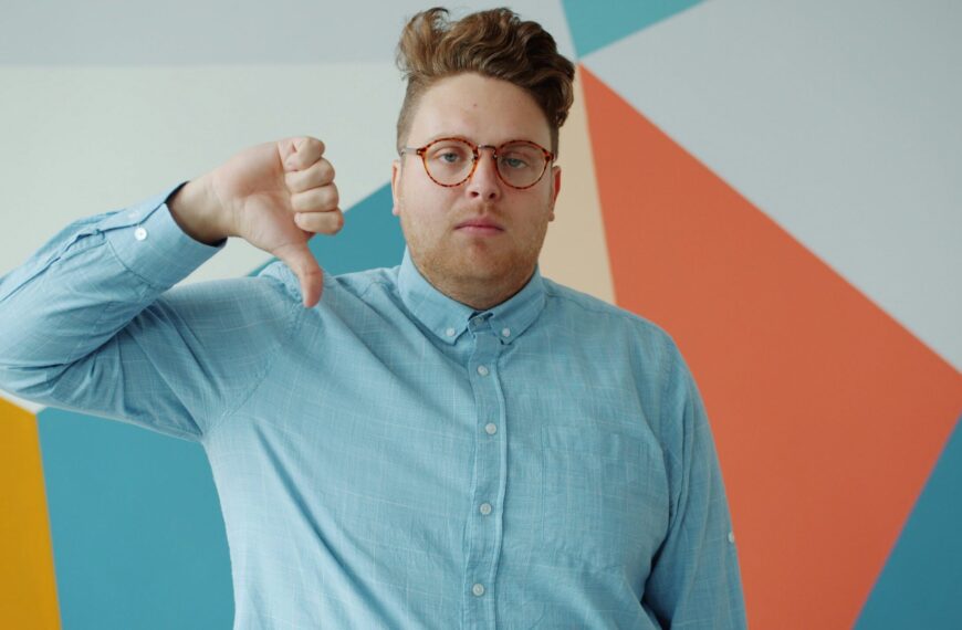 Человек в голубой рубашке показывает жест "палец вниз" на фоне геометрических фигур.