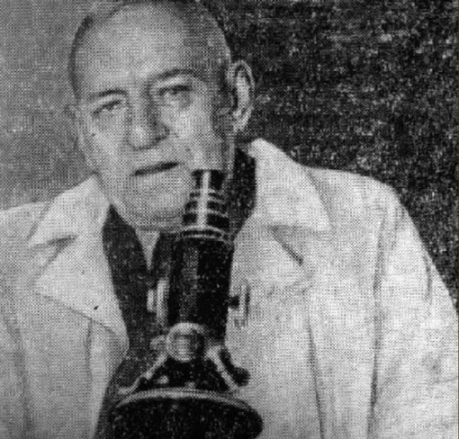 Учёный в лабораторном халате изучает объект через микроскоп, черно-белое фото.