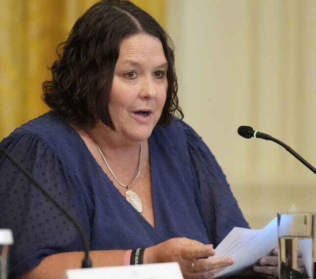 Женщина в синем платье на конференции за столом с микрофоном и документами.