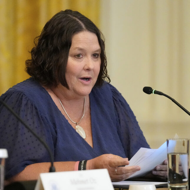 Женщина в синем платье на конференции за столом с микрофоном и документами.