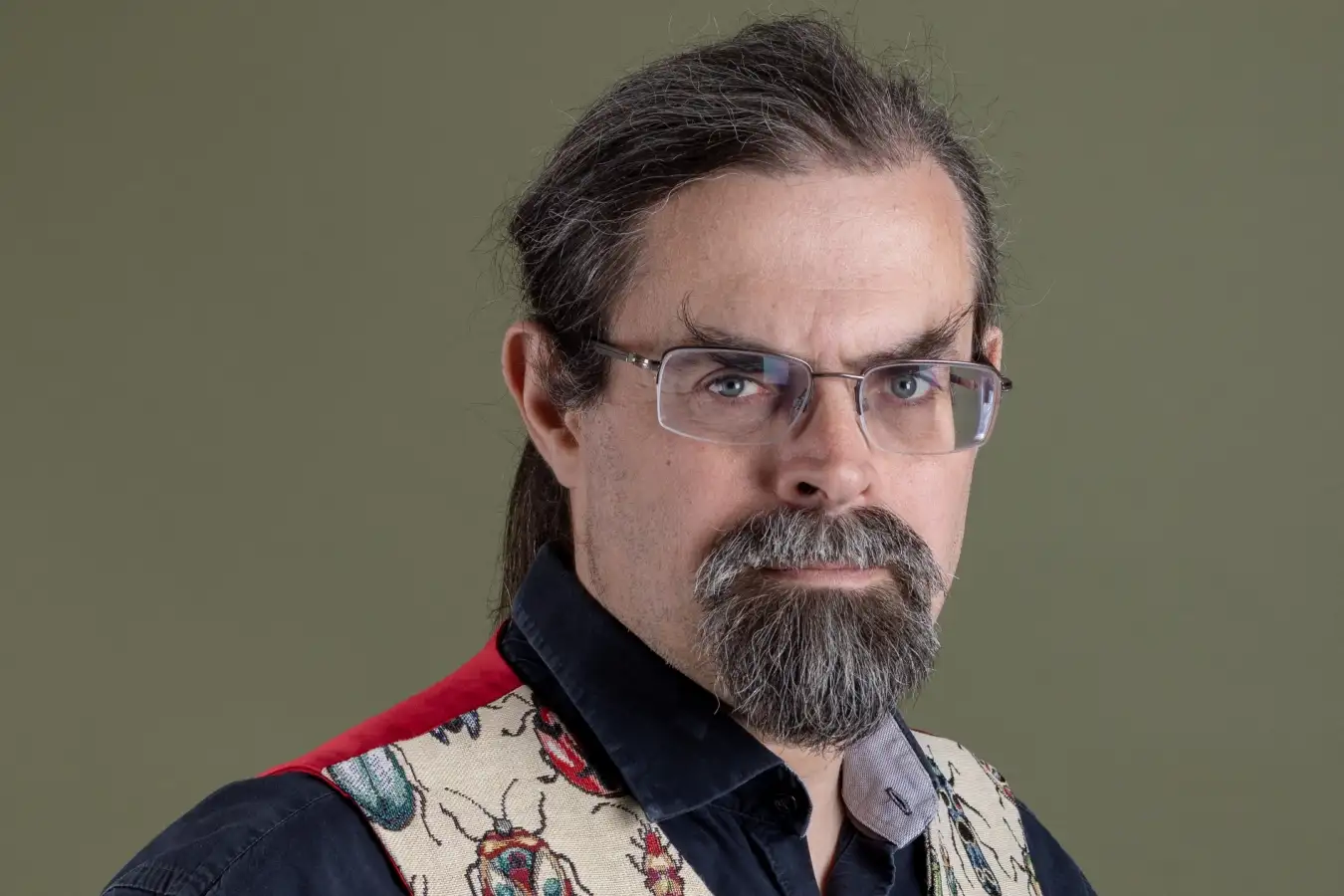 Человек в очках и жилете с жуками на фоне нейтрального цвета.