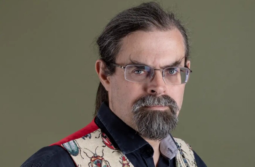 Изображение человека в очках и жилете с насекомыми на фоне оливкового цвета.