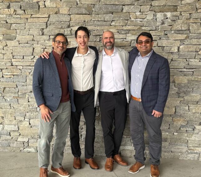 Четыре мужчины в стильной одежде стоят у каменной стены, дружеская поза.