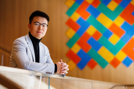 Мужчина в светлом пиджаке у стены с ярким геометрическим узором, офисная обстановка.