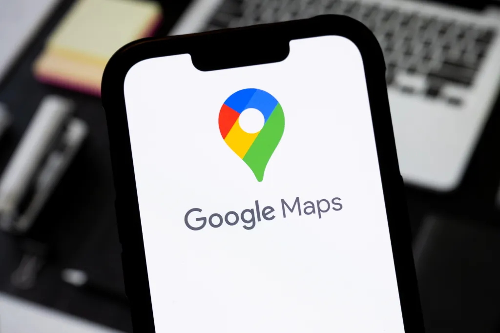 Смартфон с логотипом Google Maps на экране, фон с ноутбуком и блокнотом.