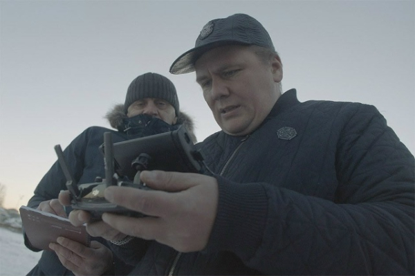 Два человека в зимней одежде управляют дроном на улице, у одного планшет в руках.