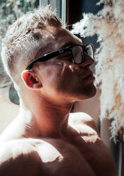 Мужчина в очках и с короткой стрижкой сидит рядом с окном и наслаждается солнечным светом.