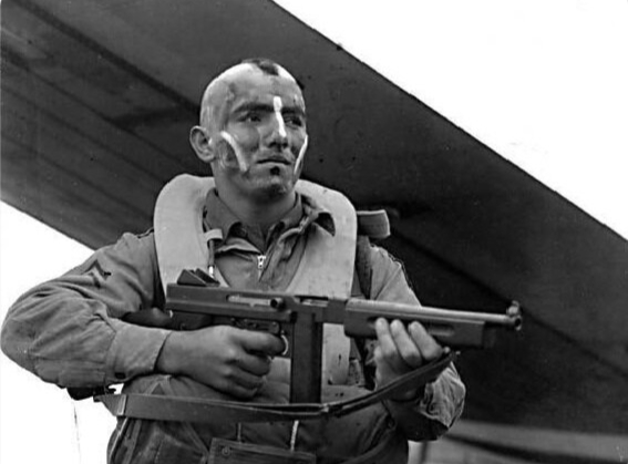 Человек в военной униформе держит автомат под крылом самолета. Историческая фотография.