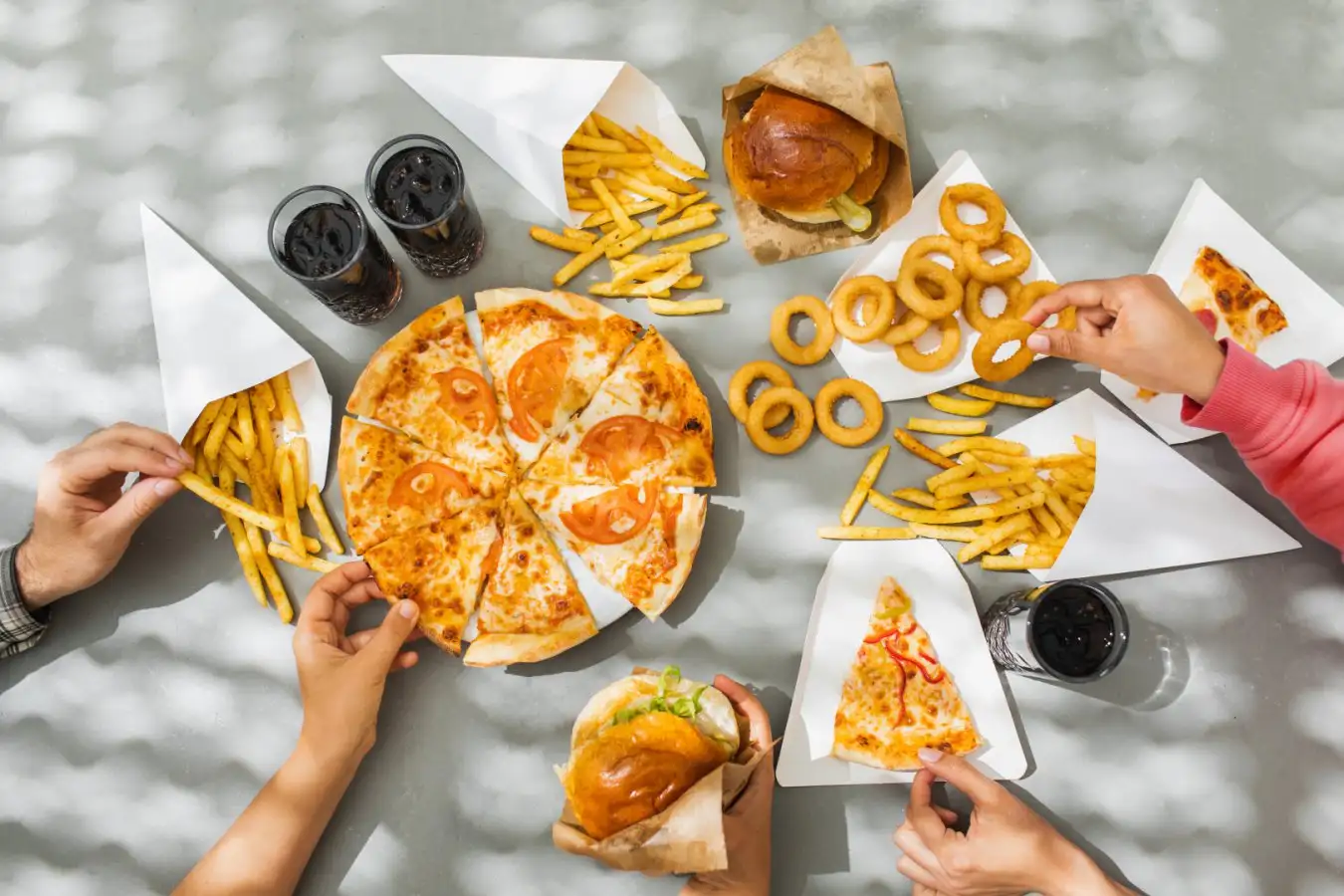 Насколько сильно следует беспокоиться о продуктах ультрапереработанной переработки? 3 Пицца, картофель фри, луковые кольца, бургер и напитки на столе, вид сверху.