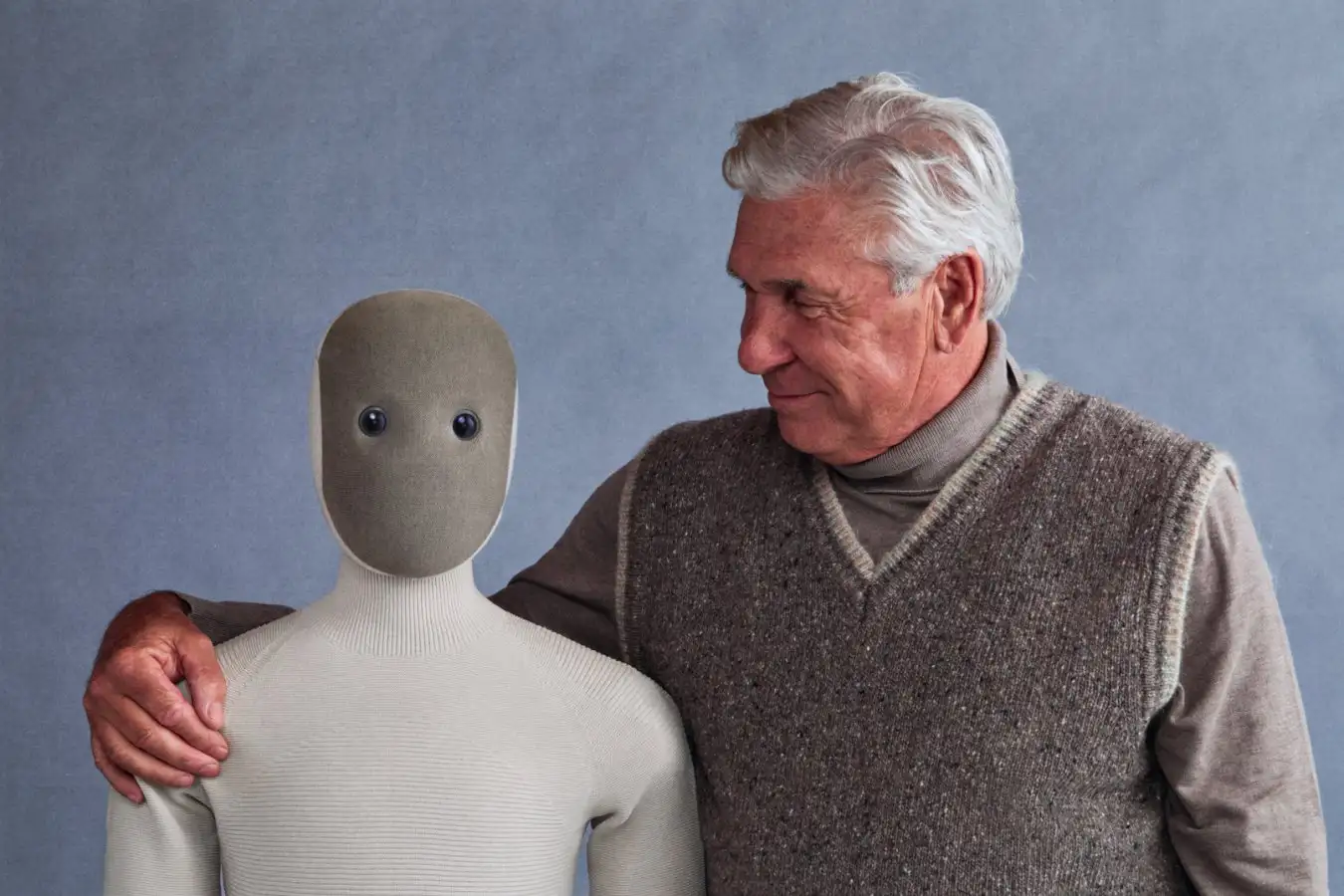 Пожилой мужчина держит манекен с кнопочными глазами на фоне серой стены.