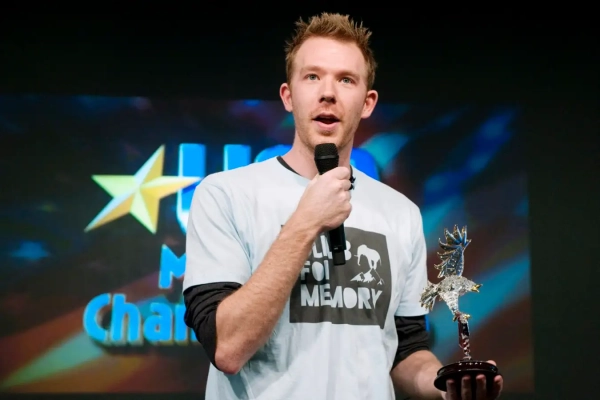 Человек в футболке с микрофоном и трофеем на сцене.