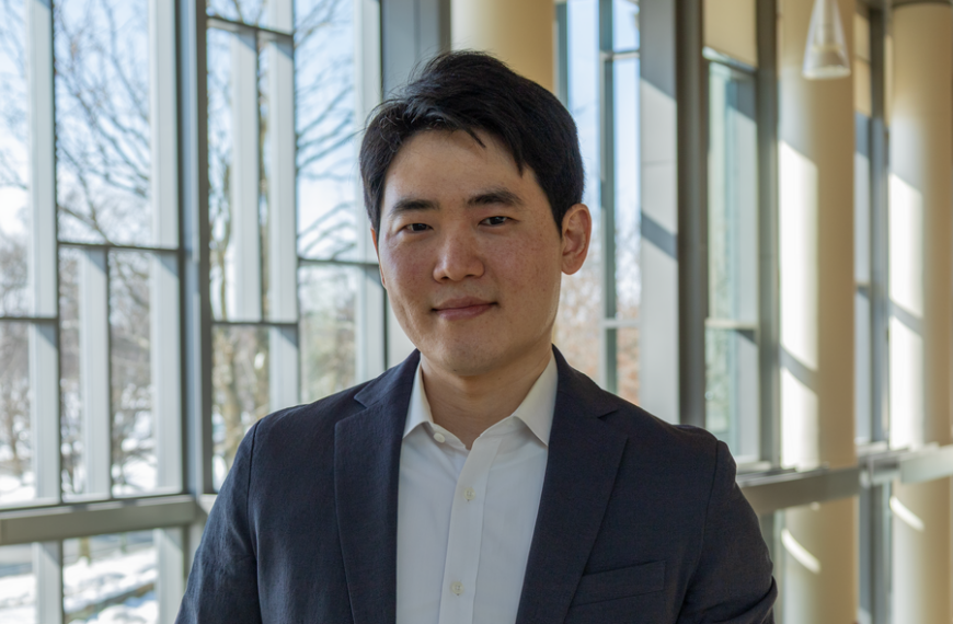 Мужчина в костюме на фоне офисного интерьера с большими окнами.