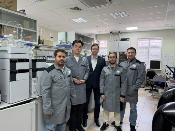 Группа людей в лаборатории, одетых в серые халаты, рядом с лабораторным оборудованием.