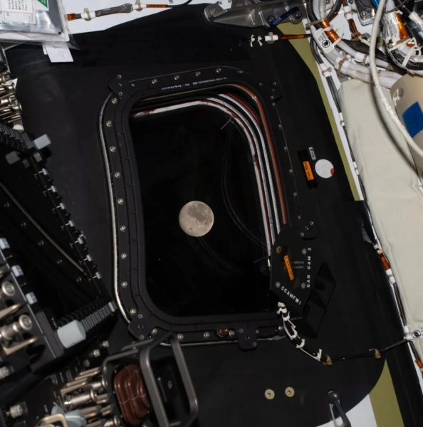 Луна видна через иллюминатор космического корабля, окруженного проводами и оборудованием.