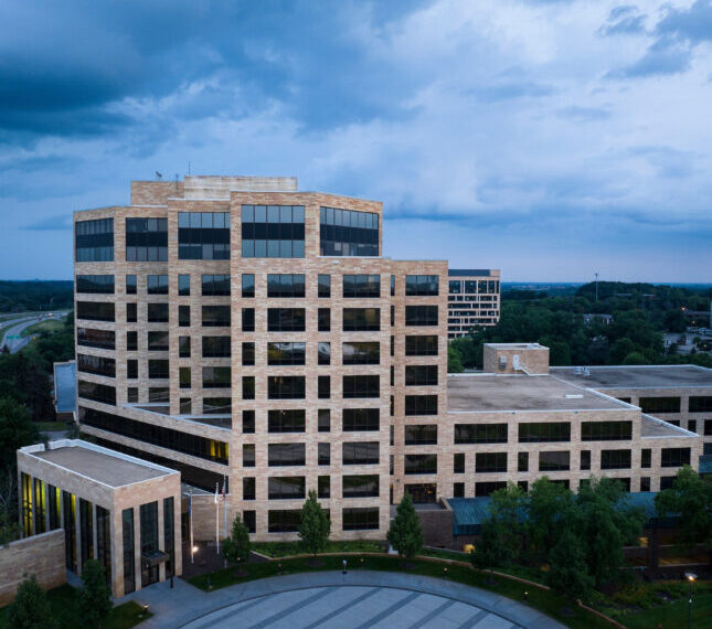 Современное офисное здание из стекла и кирпича под тучным небом, окруженное зеленью.