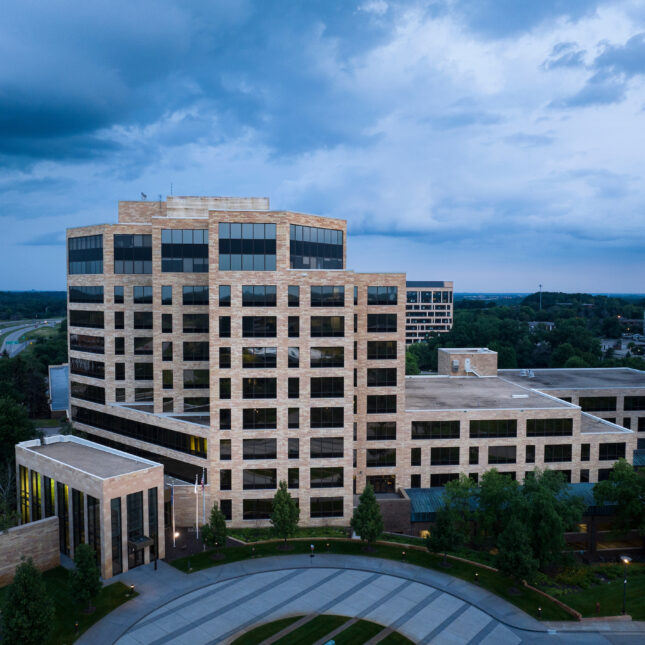 Современное офисное здание из стекла и кирпича под тучным небом, окруженное зеленью.