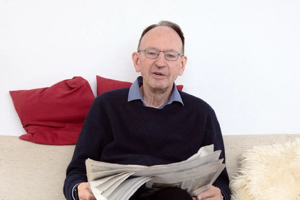 Мужчина на диване читает газету, окружён красными подушками и светлым пледом.