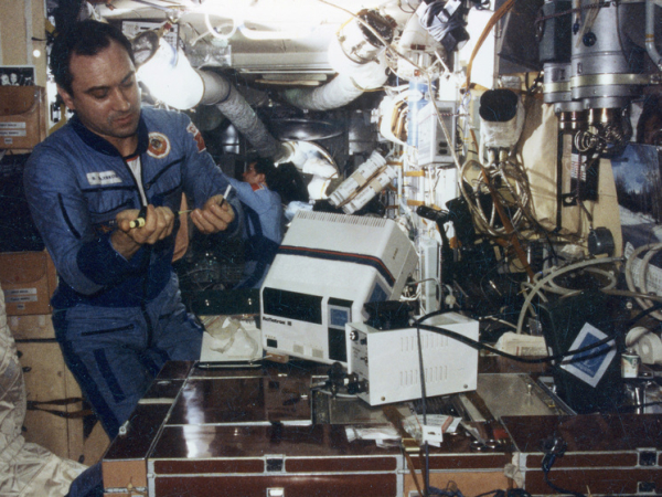 Космонавт работает на борту космической станции, окружённый оборудованием и проводами.