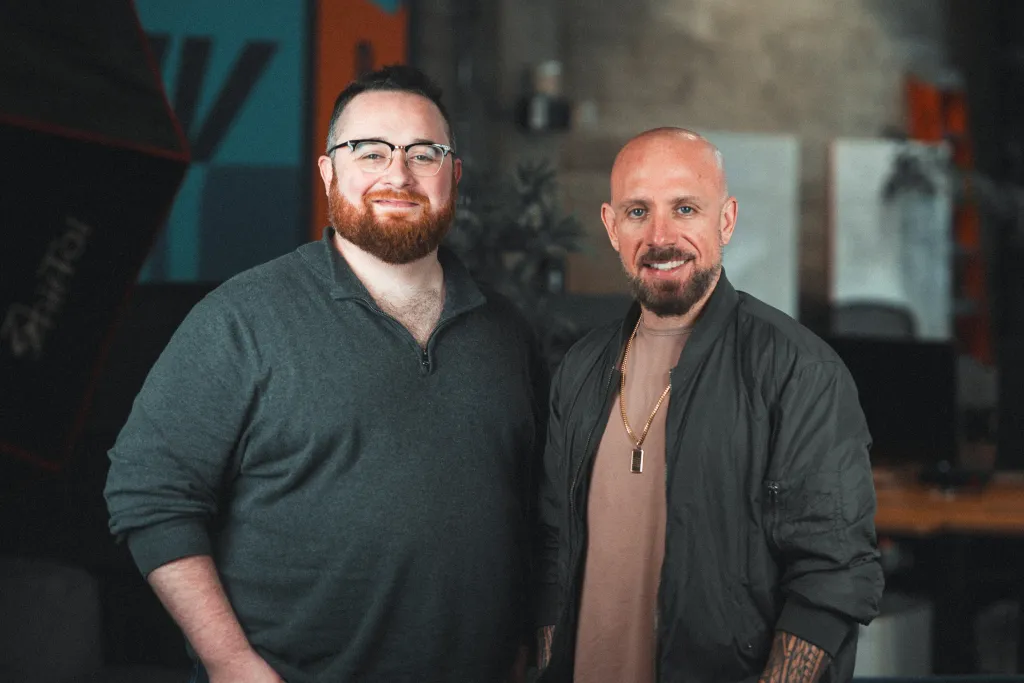 Два человека стоят рядом в помещении, один в серой кофте, другой в темной куртке с татуировками.