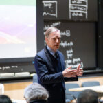 Человек выступает перед аудиторией в классе с доской и проектором на заднем плане.