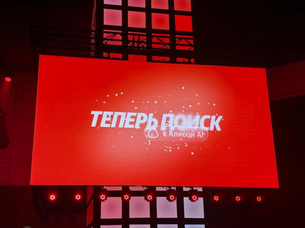 Красный экран с надписью "Теперь поиск с Алисой AI" на мероприятии.