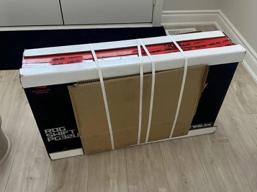 Упаковка монитора ASUS ROG Swift PG32UQ с транспортировочными ремнями на полу.