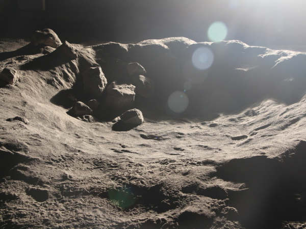 Поверхность с кратером, покрытая песком и камнями, освещена солнечным светом.