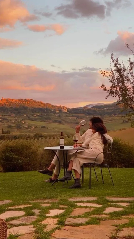 a woman sitting at a table with a bottle of wine in her hand and the sun setting behind her