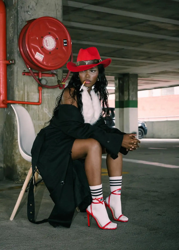 a woman sitting on top of a chair in a parking lot wearing red shoes and a hat