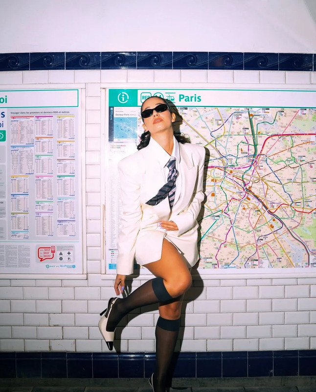 a woman posing in front of a subway map wearing stockings and knee high boots with her legs crossed